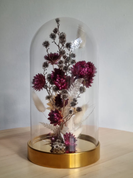 19cm glass dome with golden base featuring dried and preserved flowers in dark berry, grey and white.