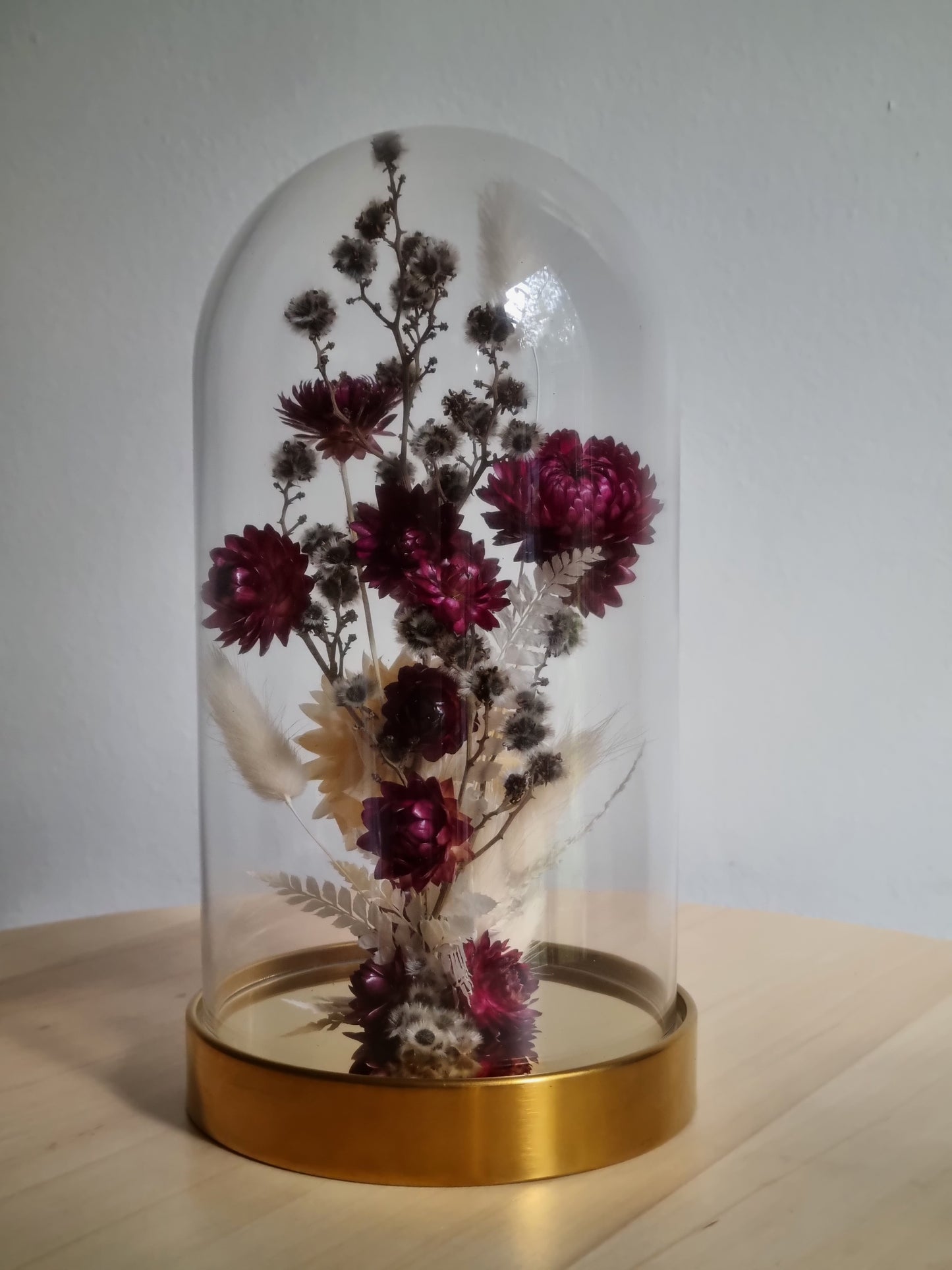 19cm glass dome with golden base featuring dried and preserved flowers in dark berry, grey and white.