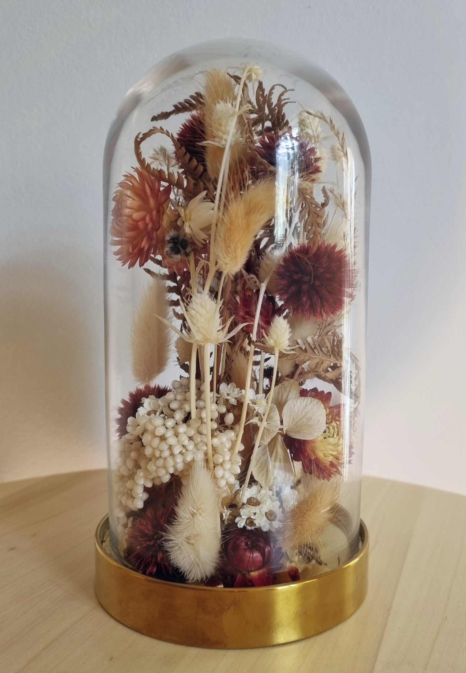 19cm glass dome with golden base features an abundance of dried wildflowers and foliages in deep reds, browns and ivory. 